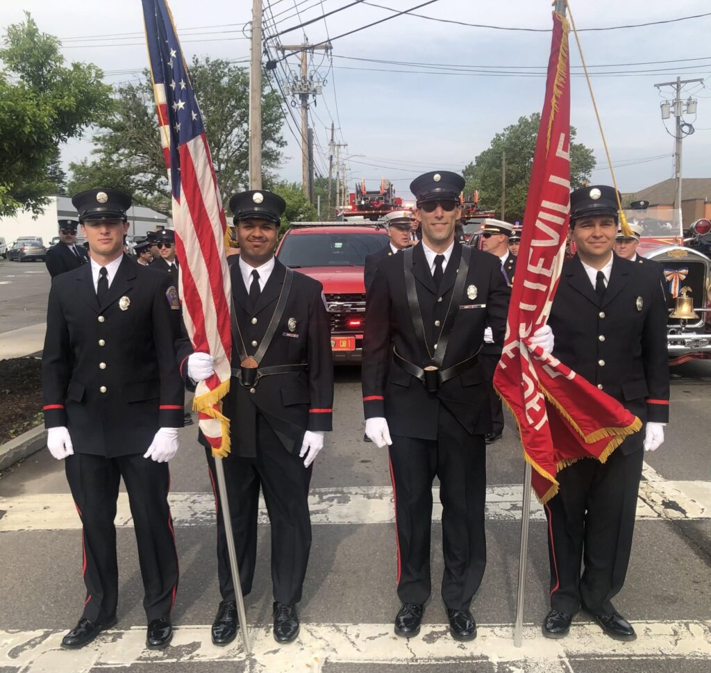 Four uniformed firefighters stand in a row on a street, two holding flags—one American flag and one red flag—while others and a red fire truck are visible in the background.