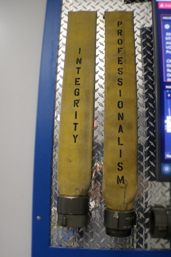 Two yellow fire hoses are mounted vertically on a wall with a diamond plate pattern. The hoses have the words "INTEGRITY" and "PROFESSIONALISM" printed on them in black, each word running down the length of a hose.