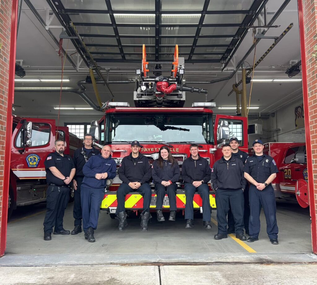 A group of ten firefighters pose in front of a fire truck inside a fire station garage, with two firefighters seated on the fire truck’s front bumper and the rest standing on either side.