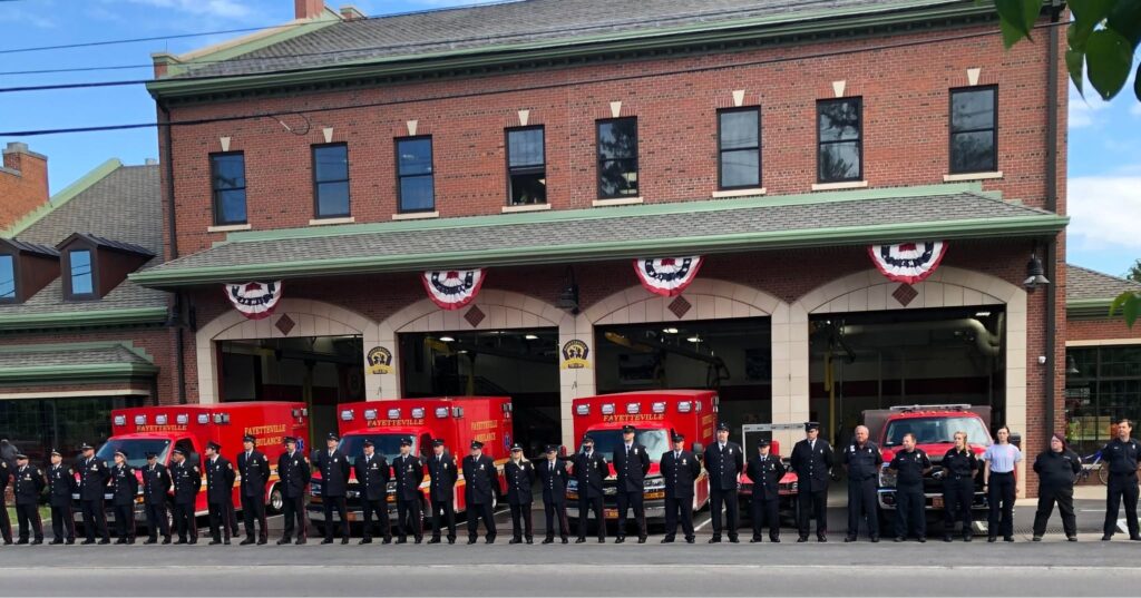 A group of firefighters in uniform stand in front of a brick fire station with three red fire trucks and patriotic bunting hanging above the garage doors.