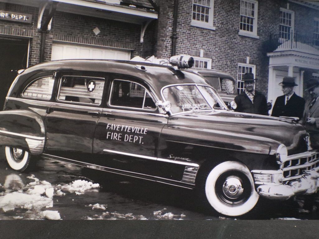 A vintage Fayetteville Fire Dept. emergency vehicle is parked outside a brick building, with a few men standing nearby. The car has whitewall tires and a roof siren. Snow is on the ground.